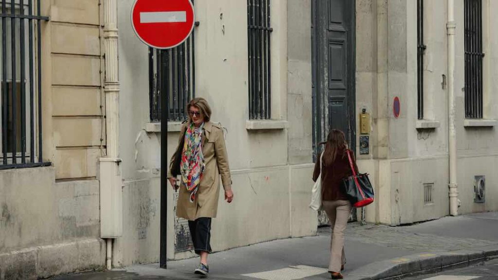 Two Parisian women crossing on the sidewalk, one wearing a trench coat and colorful scarf, the other in jeans and a light sweater